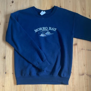 Marinblå Morro Bay sweatshirt - Säljer en marinblå sweatshirt med Morro Bay California-tryck på bröstet. Tröjan har rund hals, långa ärmar och ribbade muddar vid ärmslut och nederkant. Perfekt för chill dagar och enkel att matcha med jeans eller shorts.