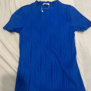 Blå ribbad topp från Amisu - Snygg blå topp från Amisu med vågigt ribbat mönster och korta ärmar. Hög hals med lätt volangkant och stretchigt material som sitter skönt. Perfekt för dig som gillar färg och vill sticka ut lite extra.