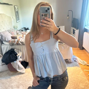 Ljusblå linnetopp med pärlknappar - Supersöt ljusblå linnetopp med breda axelband och volangig nederdel. Toppen har pärlknappar framtill och knytning i ryggen för extra detalj. Perfekt för varma dagar och ger en chill vibe till outfiten. Går att knyta en rosett bak men även bara ha banden lösa. Använd 1-2 gånger. Bra skick. Passar S-M 