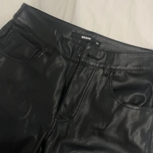 Svarta skinnbyxor från BikBok - Snygga svarta byxor i skinnimitation från BikBok. Klassisk femficksmodell med raka ben och normal passform. Byxorna har bälteshällor och dragkedja framtill. Perfekta för en edgy look och lätta att styla med allt.