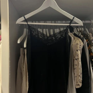 Svart spetsningar med smala axelband - Säljer en svart kortklänning med smala axelband och vacker spetsdetalj upptill. Klänningen har en elegant v-ringad hals och ärmlös design som ger en stilren och chic look. Perfekt för dig som gillar minimalistisk stil med en touch av romantik. 