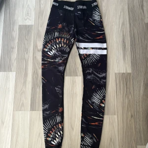Stronger Safari leggings med mönster - Snygga svarta leggings från Stronger med coolt safari-inspirerat mönster i vitt, orange och grått. Medel hög midja med elastisk logotypkant och två vita ränder på ena benet. Perfekta för träning eller chill, tillverkade i stretchigt syntetmaterial. Pris kan diskuteras. Ny pris 799kr.