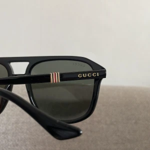 Svarta solglasögon från Gucci - Snygga svarta solglasögon från Gucci med fyrkantig form och klassisk design. Bågarna är i blank plast med guldfärgad Gucci-logga på sidorna och detaljer i rött och vitt. Perfekta för dig som vill ha en stilren och lyxig look.
