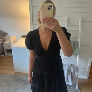 Svart omlottklänning med puffärm - Säljer en svart kortklänning med omlott och djup v-ringning. Klänningen har puffiga kortärmar och markerad midja, samt volanger i kjolen. Materialet är lätt och har diskreta spetsdetaljer som ger en extra touch. Perfekt för dig som gillar en feminin och trendig look.