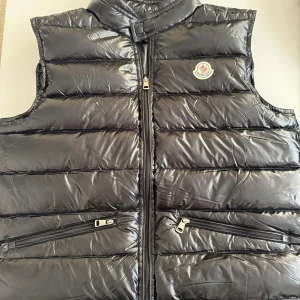 Moncler väst - Snygg svart dunväst från Moncler med glansig finish och klassisk logga på bröstet. Västen har två praktiska fickor med dragkedja och hög krage med tryckknapp. Perfekt för lager-på-lager och streetstyle.