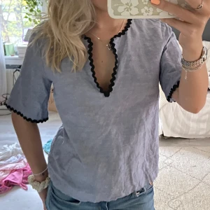 Ljusblå blus med svarta vågiga kanter från & Other Stories - Säljer en ljusblå blus från & other Stories med unika svarta vågiga kanter längs v-ringningen och ärmsluten. Blusen har korta ärmar och är tillverkad i ett lätt linnematerial. Perfekt för vår och sommar!