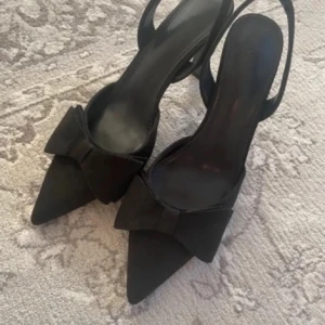 Svarta pumps med rosett och spetsig tå - Säljer ett par eleganta svarta pumps med spetsig tå och stor rosett framtill. Skorna har öppen häl med rem och en klassisk, stilren design som passar perfekt till många outfits.