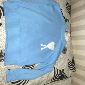 Ljusblå stickad tröja från Ami Paris - Säljer en ljusblå stickad tröja från Ami Paris med det ikoniska vita hjärta-A-motivet på bröstet. Tröjan har rund halsringning, långa ärmar och breda ribbade muddar vid ärmslut och nederkant. Perfekt för dig som gillar stilrena och trendiga plagg.