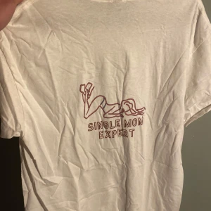 Vit t-shirt med tryck 'Single Mon Expert' - Vit t-shirt med ett minimalistiskt neon tryck av en person och texten 'Single Mon Expert' på bröstet. Klassisk passform och korta ärmar. Perfekt för dig som gillar unika och roliga prints. Helt ny har bara legat