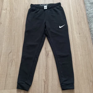 Svarta mjukisbyxor från Nike - Svarta mjukisbyxor från Nike med Dri-FIT-material och elastisk midja. Byxorna har en smal passform och klassisk Nike-logga på ena benet. Perfekta för träning eller chill. Säljes i storlek S.