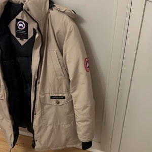 Beige parkajacka från Canada Goose - 🇸🇪Säljer en beige parkajacka från Canada Goose med svart innerfoder och klassisk logga på ärmen. Jackan har huva, dragkedja och knappar framtill samt flera fickor. Perfekt för kyliga dagar.🇫🇮Beige Canda Goose takki, Käytetty.