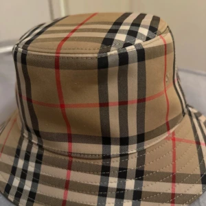Rutig bucket hat från Burberry - Säljer en klassisk bucket hat från Burberry i deras ikoniska beigea rutiga mönster med svarta, vita och röda ränder. Hatten har bred brätte och är tillverkad i bomull. Perfekt accessoar för att lyfta din stil. 8y-12y storlek 