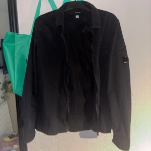 Svart overshirt från C.P. Company - Säljer en svart overshirt från C.P. Company med dragkedja framtill och klassisk linsdetalj på ärmen. Jackan har en enkel, stilren design och lång ärm. Perfekt för lager-på-lager och streetwear.