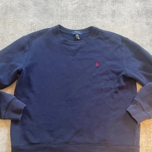 Mörkblå sweatshirt från Polo Ralph Lauren - Klassisk mörkblå sweatshirt från Polo Ralph Lauren med rund halsringning och diskret röd logga broderad på bröstet. Tröjan har långa ärmar och ribbade muddar vid ärmslut och nederkant. Perfekt för en avslappnad stil.