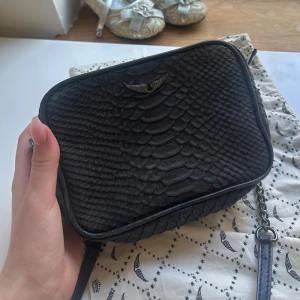 Svart Zadig&Voltaire  i ormskinnsmönster - Säljer en svart Zadig väska med ormskinnsmönster och svartfärgad detalj framtill. Väskan har dragkedja upptill och en axelrem. 