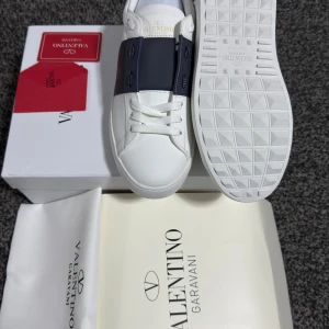 Valentino Garavani vita sneakers med mörkblå detalj - Snygga vita sneakers från Valentino Garavani med en bred mörkblå rem över snörningen. Skorna har klassisk rund tå och platt vit sula. Perfekta för dig som vill ha en stilren och exklusiv look.