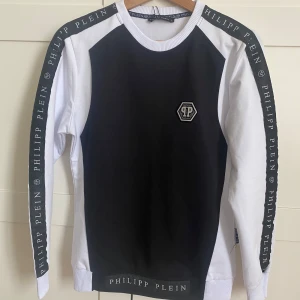 Svart och vit tröja från Philipp Plein - Låmgärmad tröja från Philipp Plein med logga på bröstet och text längs ärmarna och nederkanten. Tröjan har rund halsringning och ribbade muddar. Aldrig använd.