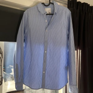 Blåvit randig skjorta från 157 - Klassisk blå och vit randig skjorta från 157 med lång ärm och knappar framtill. Skjortan har en tidlös design med smal krage och passar perfekt till både jeans och chinos. Tillverkad i mjuk bomull för en bekväm känsla.