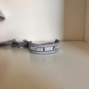 Blått armband från Dior - Säljer ett snyggt blått armband från Dior med texten 'Christian Dior J'Adior' broderat. Armbandet har justerbar knytning med tofsar i ändarna. Perfekt accessoar för att lyfta din stil.