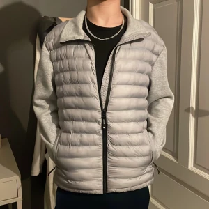 Grå dunväst med ärmar från Superdry - Snygg grå dunväst med ärmar från Superdry. Västen har en dragkedja framtill och är quiltad för extra värme. Perfekt för kyliga dagar med sin stilrena design och bekväma passform. Knappat använd och den passar 164-170