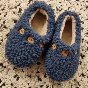 Blå fluffiga tofflor med björnmotiv - Mysiga blå tofflor med fluffig yta och små broderade björnar på ovansidan. Perfekta för att hålla fötterna varma hemma. Insidan är mjuk och skön mot foten.