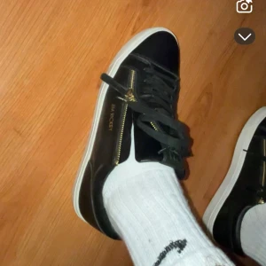 Svarta sneakers från Jim Rickey med gulddragkedja - Säljer ett par svarta sneakers från Jim Rickey med guldfärgade dragkedjor på sidorna och vita sulor. Skorna har snörning och en stilren design med märkets logga i guld på sidan. Perfekta för dig som gillar en clean och modern look. Kan tänka mig byta mot andra saker. De är köpta för 2800kr använda ca 10 gånger men fortfarande nya. 
