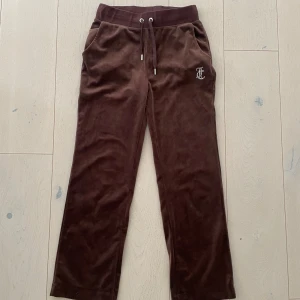 Bruna velourbyxor från Juicy Couture - Mysiga bruna velourbyxor från Juicy Couture med dragsko i midjan och fickor. Glittrig JC-logga framtill och glittrig 'Juicy' text bak. Perfekta för en avslappnad och trendig look.