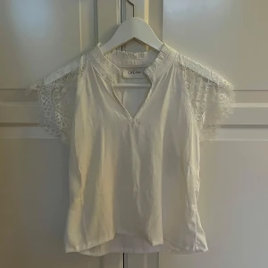 Vit blus med spetsdetaljer från Cream - Säljer en vit blus från Cream med v-ringning och vackra spetsdetaljer på ärmarna. Blusen har korta, luftiga ärmar i spets och en mjuk, feminin design. Perfekt för en fräsch och stilren look.