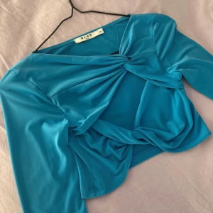 Turkos långärmad croppad topp från NA-KD - Säljer en turkos långärmad croppad topp från NA-KD med twistad knutdetalj framtill. Toppen har en snygg och unik design som passar perfekt till jeans eller kjol. Materialet är mjukt och stretchigt för en bekväm passform.