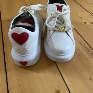 Vita sneakers från Love Moschino  - Säljer ett par vita sneakers från Love Moschino med röda hjärtan på hälen och sulan samt guldfärgad hjärtdetalj vid snörningen. Skorna har tjock vit sula och är perfekta för dig som gillar unika detaljer.
