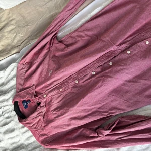 Vinröd skjorta från GANT - Säljer en klassisk rosa skjorta från GANT i storlek M. Skjortan har lång ärm, knappar framtill och en liten broderad logga på bröstet. Perfekt för dig som gillar stilrena och tidlösa plagg.