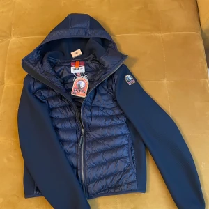 Parajumpers jacka - Säljer en mörkblå hybridjacka från Parajumpers med quiltad front och rygg, samt släta ärmar. Jackan har huva, dragkedja och en snygg logotyp på ärmen. Perfekt för dig som vill ha bekväm jacka som samtidigt ser bra ut.                        Säljs pågrund av att jag inte riktigt använder den längre då jag har köpt nya jackor! Jackan är verkligen felfri, vid snabb affär kan jag tänka mig sänka priset lite!💯