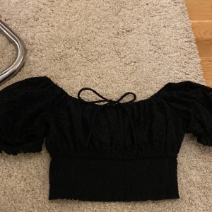 Svart croppad blus från FB Sister - Supersöt svart croppad blus från FB Sister med puffärmar och knytning framtill. Blusen har broderade hål som ger en fin struktur och offshoulder-modell. Perfekt för dig som gillar trendiga detaljer och en feminin look.