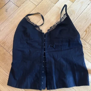 Svart spetsdetaljerad korsett-topp - Säljer en svart korsett-topp med smala axelband och fina spetsdetaljer längs urringningen. Toppen har knäppning framtill och en figurnära passform som framhäver siluetten. Perfekt för att lyfta vilken outfit som helst.