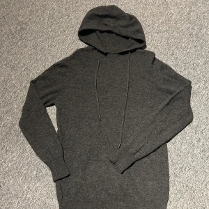 Merinoull hoodie  - Mysig mörkgrå hoodie med huva och dragsko. Tröjan är stickad och har långa ärmar. Perfekt för kyliga dagar och enkel att matcha med det mesta. Ett litet hål i ena armen som knappt syns men har med de ändå 