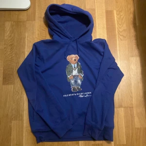 Blå hoodie med björn från Polo Ralph Lauren - Säljer en blå hoodie från Polo Ralph Lauren med den klassiska Polo Bear-trycket på bröstet. Tröjan har huva och ribbade muddar vid ärmslut och nederkant. Perfekt för en avslappnad och stilren look. Köpt för 2500kr för ca ett halvår sedan.