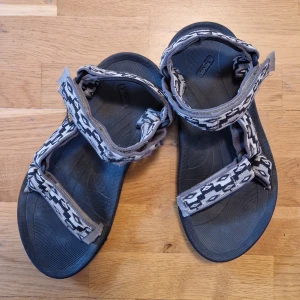 Svart och gråa sandaler från Teva - Klassiska Teva sandaler. Perfekta för sommarens utomhusaktiviteter - justerbara både i fram och runt anklarna, bekväma att gå i och tål att bli blöta.