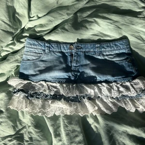 Jeanskjol med volang och spets - Supersöt kort jeanskjol med volangkant i vit broderad spets och blåvitrandigt tyg. Midjemått 42cm och längd 28! OBS!!! Man behöver oftast större midjemått i kjolar då man oftast har den längre ner på höfterna. Jag har midjemått 38-39cm i jeans.