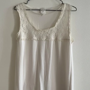 Vit ärmlös topp med spets från Monki - Supersöt vit ärmlös topp från Monki med spetsdetaljer upptill och längs nederkanten. Toppen har rund halsringning och är tillverkad i mjukt material med fina spetsinslag som ger en romantisk känsla. Finns en svag obehandlad fläck, se bild