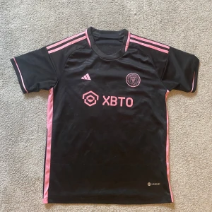Inter Miami CF Messi 10 fotbollströja - Säljer en svart och rosa Inter Miami CF fotbollströja från Adidas med Messi 10 på ryggen. Tröjan har korta ärmar, rosa detaljer och klubbmärke på bröstet. Perfekt för dig som älskar fotboll och Messi!