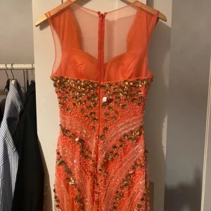 Glittrig långklänning i orange/rosa med pärlor och paljetter - Säljer en fantastisk orange långklänning med transparent axelparti. Klänningen är dekorerad med pärlor och paljetter i guld och grönt Perfekt för bal eller festliga tillfällen. Väldigt bra skick där uppe men sliten där nere vissa pärlor ramlar av och vissa trådar sticker ur därav de billiga priset. 
