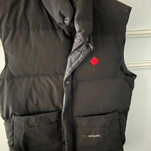 Svart dunväst från Canada Goose - Säljer en svart dunväst från Canada Goose med klassisk design. Västen har två stora fickor framtill, diskret logga med röd lönnlöv på bröstet och hög krage. Hör av er om ni undrar något 