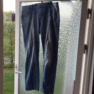 Blå jeansbyxor från Firetrap - Säljer ett par klassiska blå jeansbyxor från Firetrap med raka ben och slitna detaljer framtill. Byxorna har fem fickor och normal passform. Perfekta för dig som gillar en avslappnad stil.