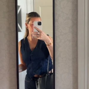 Mörkblå ärmlös denimväst med knappar - Snygg mörkblå denimväst med v-ringning och knäppning framtill. Västen har en klassisk passform och är perfekt att styla över en t-shirt eller blus. Enkel och stilren design som passar till många outfits. Normal till liten i storlek. Jag är en 38/40!