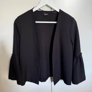 Ny Svart kavajjacka med volangärm från Ginatricot - Stilren svart kavajjacka från Ginatricot med öppen front och markerade volangärmar. Jackan har en rak passform och är tillverkad i ett mjukt, slätt material. Perfekt för att lyfta en enkel outfit.