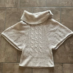 Beige stickad tröja med polokrage - Säljer en beige stickad tröja med bred polokrage som kan knäppas med en knapp. Tröjan har korta fladdermusärmar och ett snyggt kabelstickat mönster framtill. 