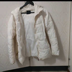 Vit pufferjacka med huva från LINDEX - Säljer en vit pufferjacka från LINDEX med huva och fuskpälsdetalj. Jackan har dragkedja framtill och två fickor med dragkedja. Perfekt för kyliga dagar och har en klassisk, enkel design.