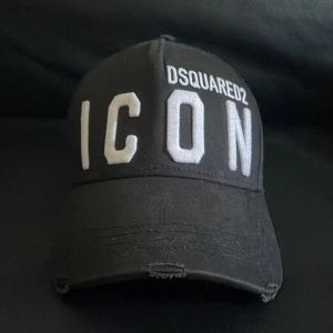 Svart keps från Dsquared2 med broderad text - Svart keps från Dsquared2 med stor vit broderad text 'ICON' framtill och 'DEAN & DAN CATEN' baktill. Justerbart spänne bak för perfekt passform. Klassisk böjd skärm och ventilerande hål upptill.