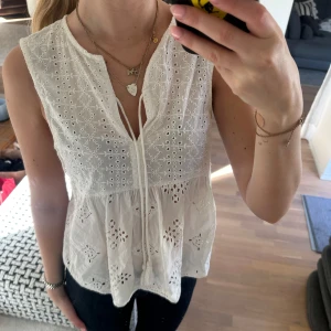 Vit ärmlös blus med broderade detaljer - Säljer en söt vit ärmlös blus med broderade blommönster och spetsdetaljer. Blusen har v-ringning med knyt framtill och en luftig, lätt utsvängd passform. 