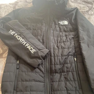 Svart quiltad jacka från The North Face - Säljer en svart quiltad jacka från The North Face med huva och dragkedja framtill. Jackan har två sidofickor med dragkedja och logga på bröstet samt ärmen. Perfekt för kyliga dagar.